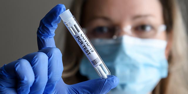 Woman wearing a light blue mask holding a Covid-19 PCR test tube in a dark blue latex glove