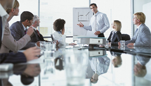 Business people of varying genders and ethnicities gathered at a conference table watching a presentation